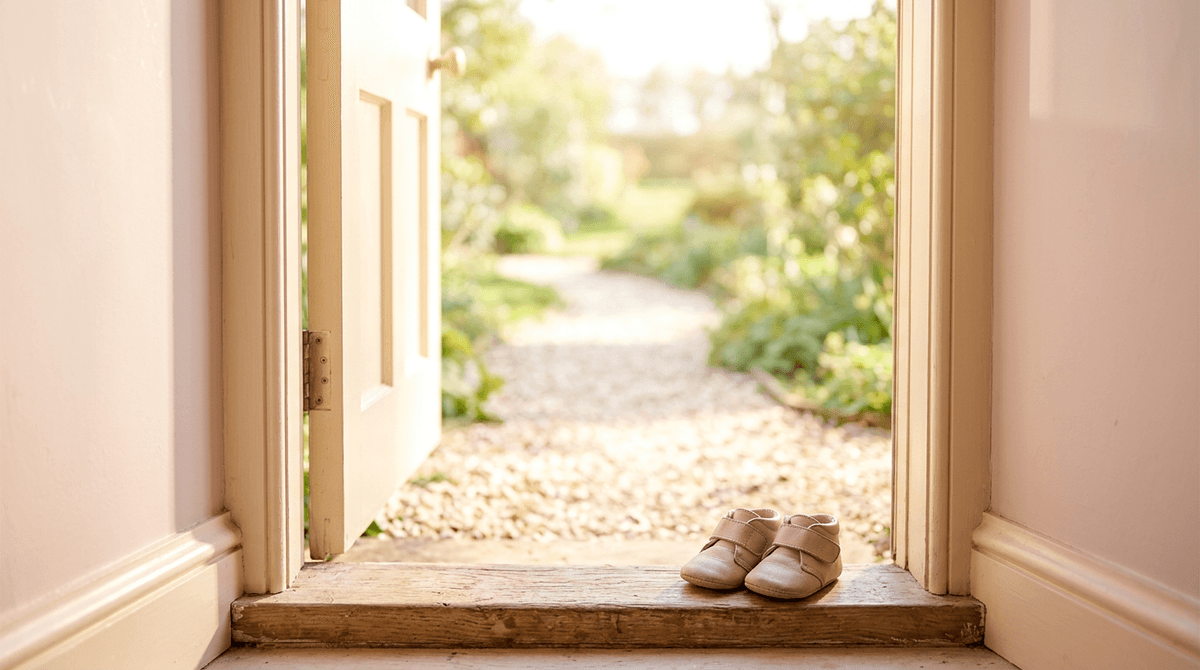 A sunlit path visible through an open doorway with baby shoes on the sill, evoking an optimistic forward look