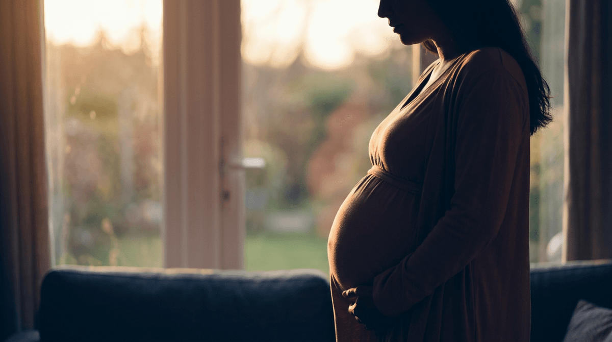 Silhouette of a pregnant woman standing near a window, backlit by golden hour light